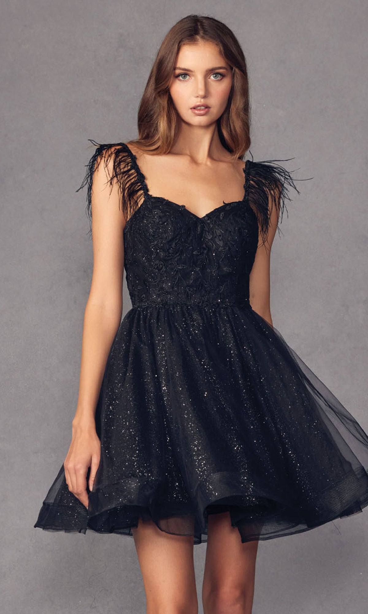 A woman wears the Short Babydoll Homecoming Dress with Feathers 881, featuring glitter, feathered straps, an embroidered sweetheart bodice, and a tulle skirt, standing before a plain gray background.